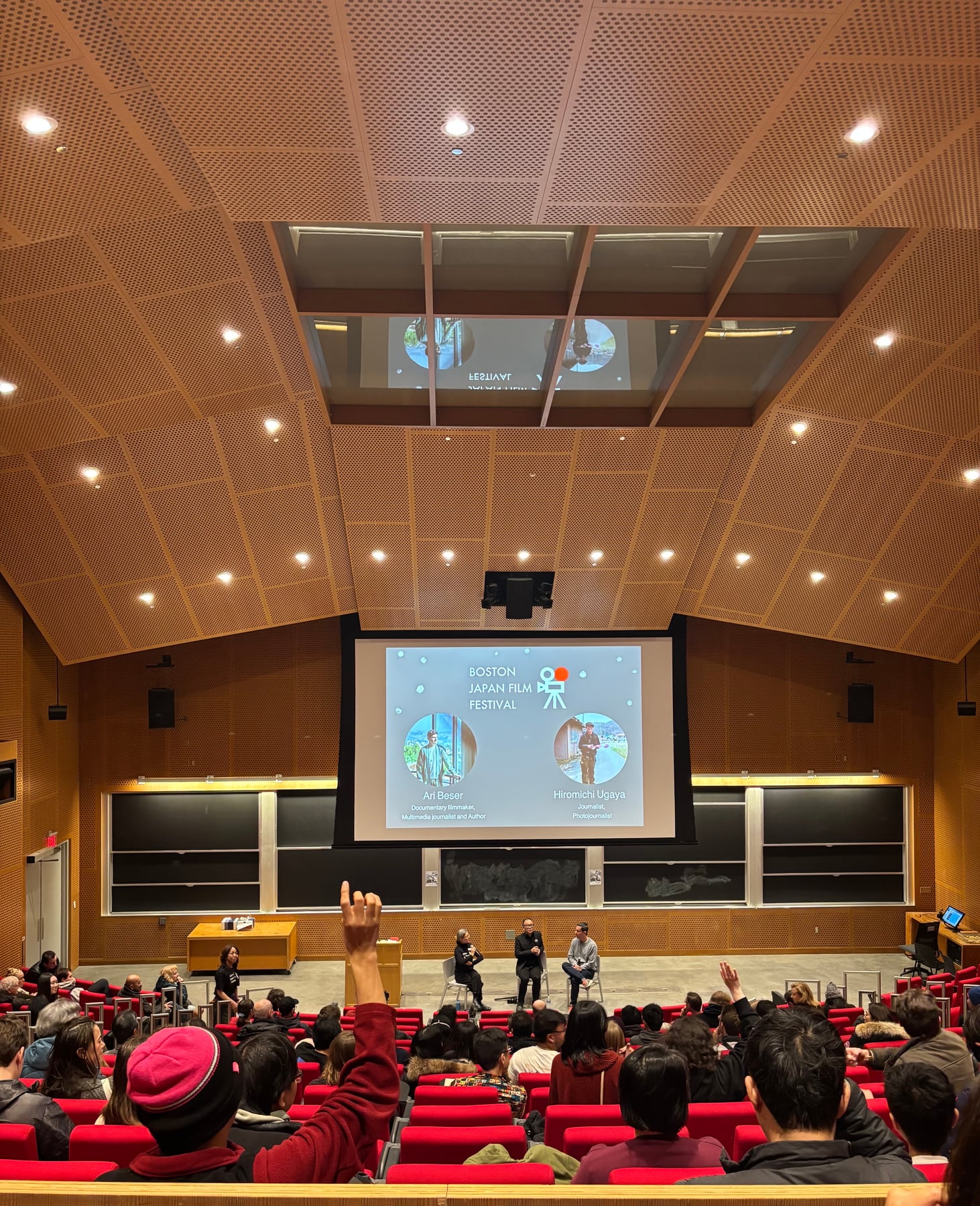 A view from the back of the Boston Japan Film Festival auditorium, with raised hands in the audience during a discussion.
